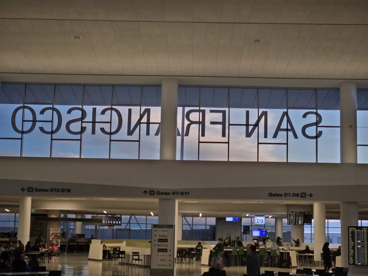 An airport terminal interior with large windows displaying the reversed text 'san francisco' against a twilight sky. Below…