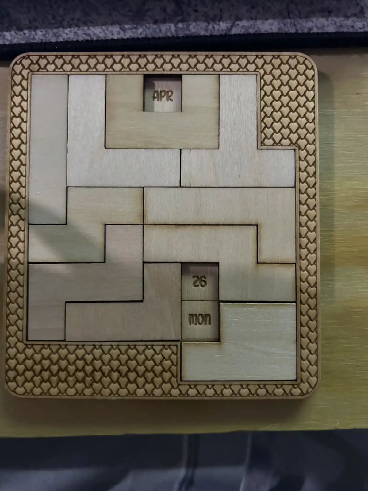 A wooden puzzle calendar with interlocking pieces, showing 'APR' at the top and '26' and 'MON' at the bottom, indicating…