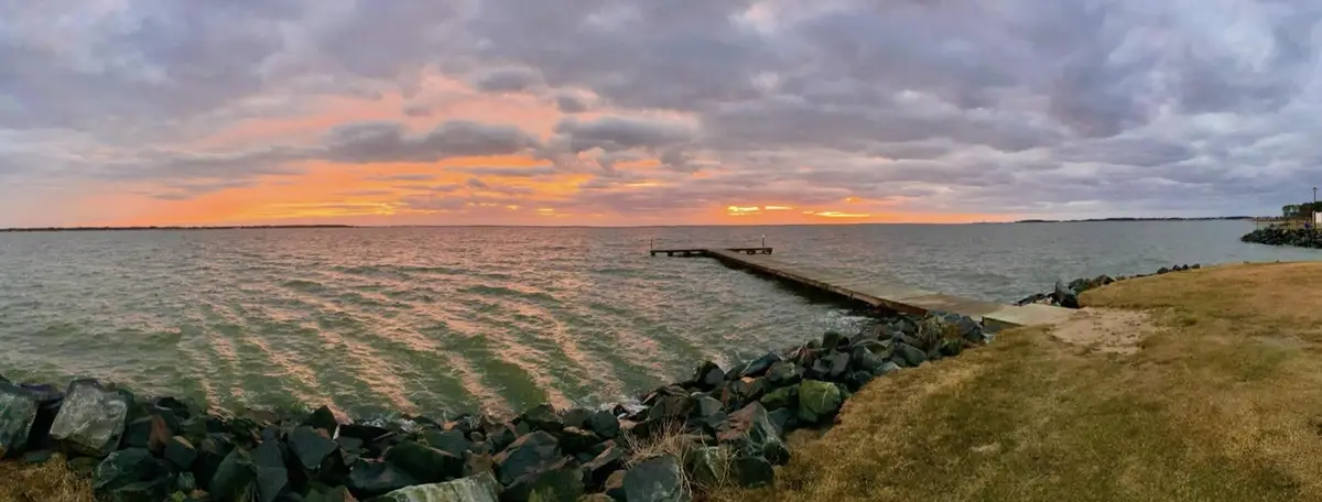 A scenic photo of a wooden pier extending into a body of water during a colorful sunset with orange and pink clouds. The…