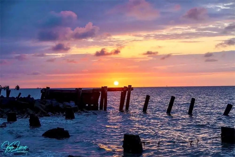 A serene sunset over the ocean with a decaying wooden pier structure in the foreground, its posts partially submerged in…