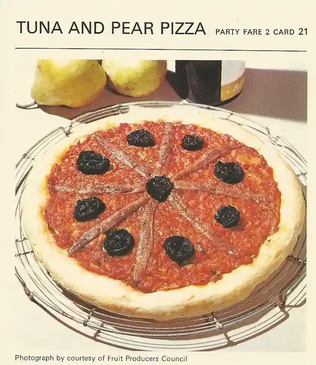 A vintage recipe card image shows a pizza with tomato sauce, arranged slices of canned tuna, and dark, round pears as…