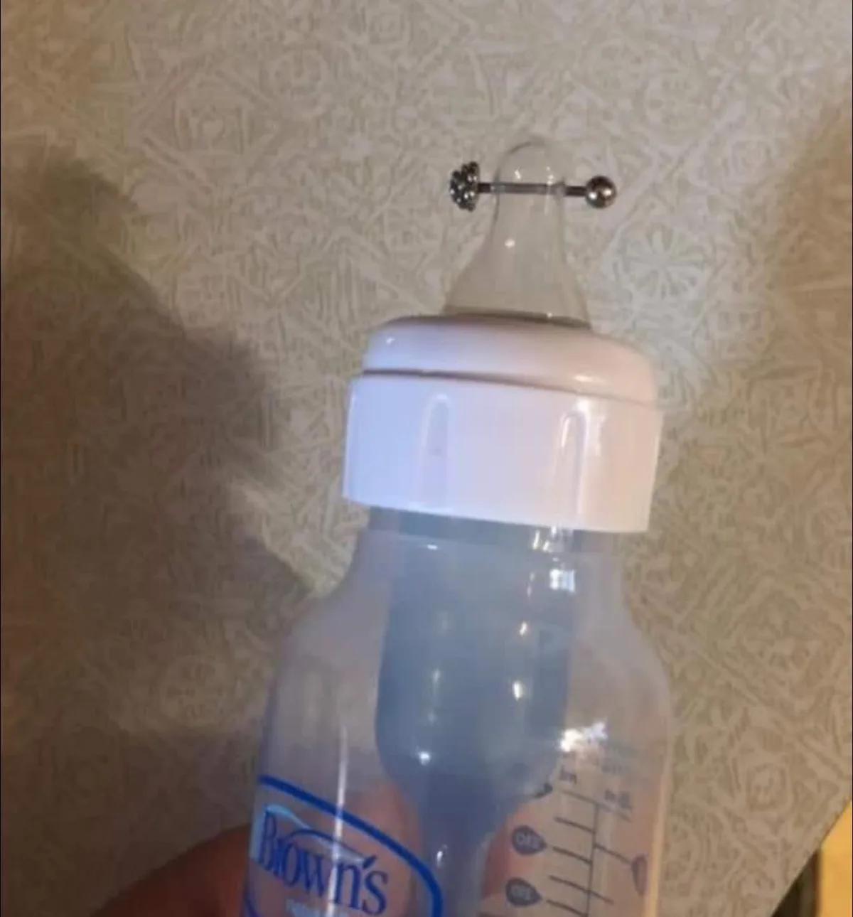 A close-up photo shows a clear plastic baby bottle with a white cap, held against a patterned wallpaper background. The…