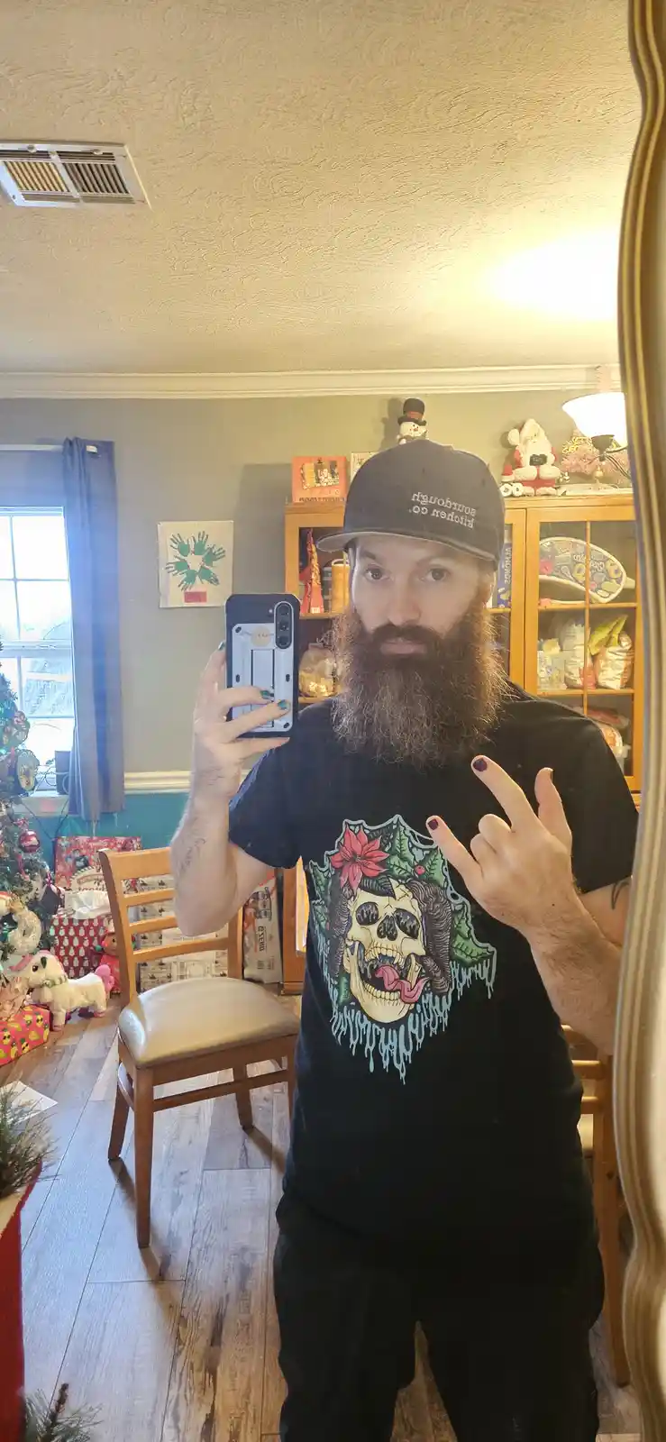 A man with a long beard and a black t-shirt featuring a skull with a poinsettia and holly takes a mirror selfie in a…