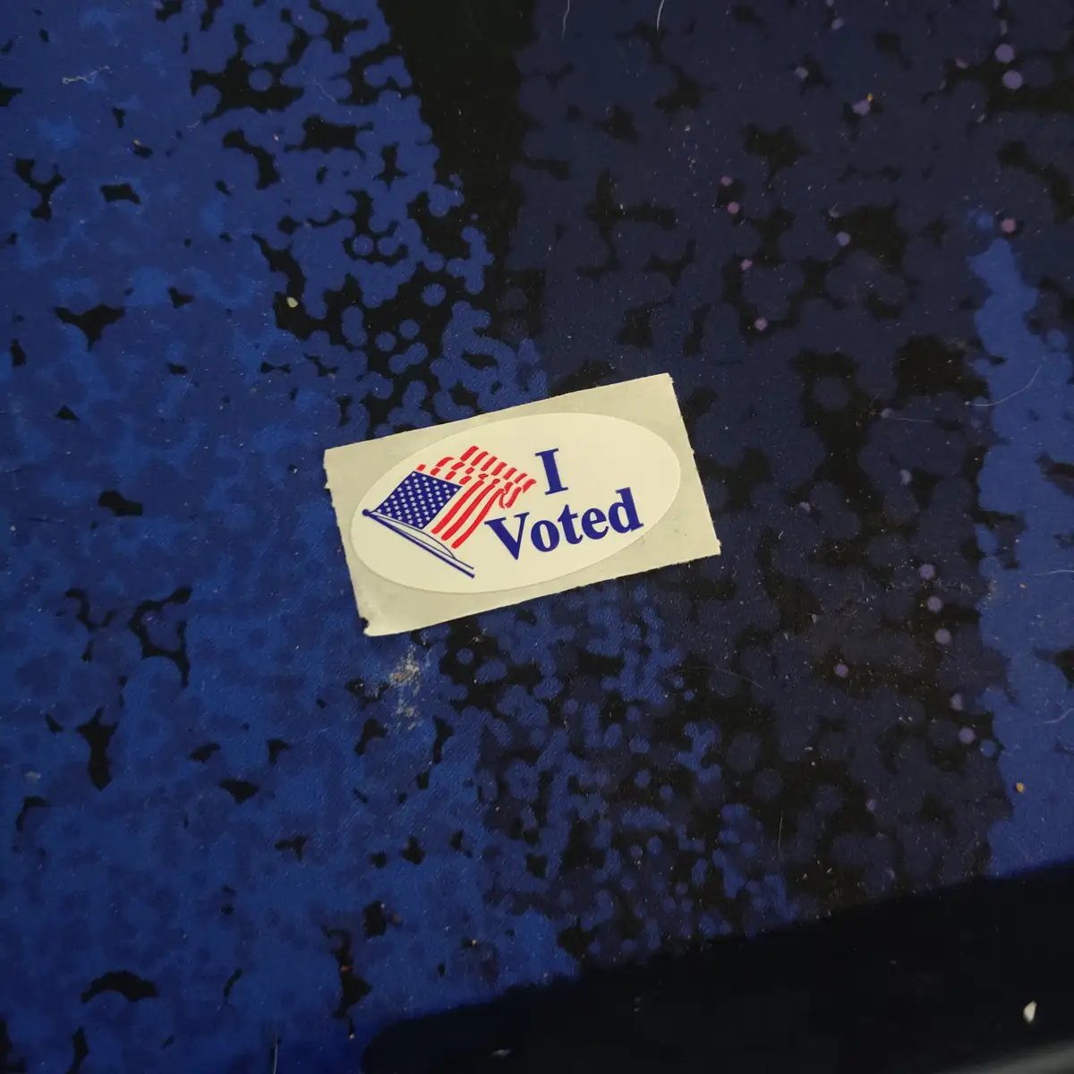 A close-up photo shows a standard American 'I Voted' sticker with a flag graphic, lying on a speckled blue and black…