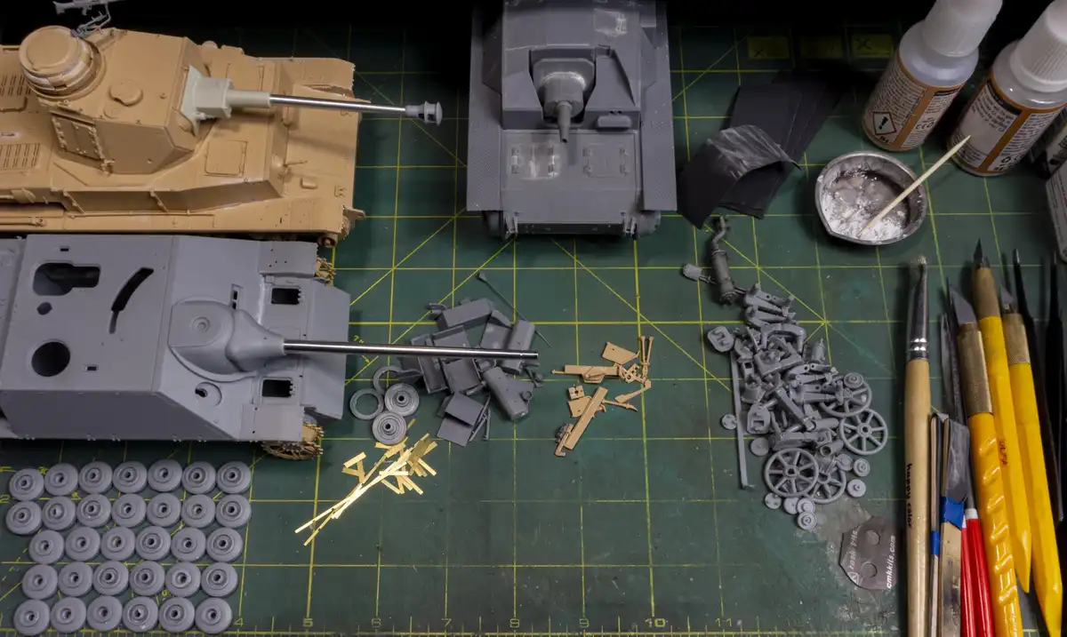 An overhead shot of a model-building workspace with three partially assembled tanks, scattered plastic parts, glue bottles…