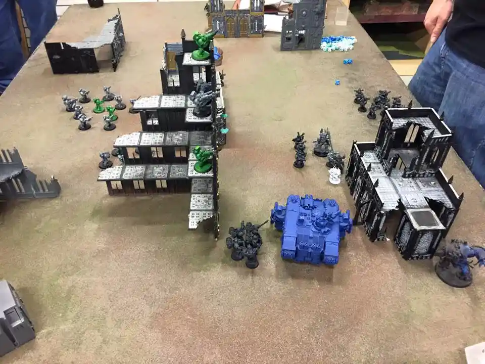A tabletop wargaming scene shows miniature figures and buildings on a brown surface; blue-armored miniatures and a large…