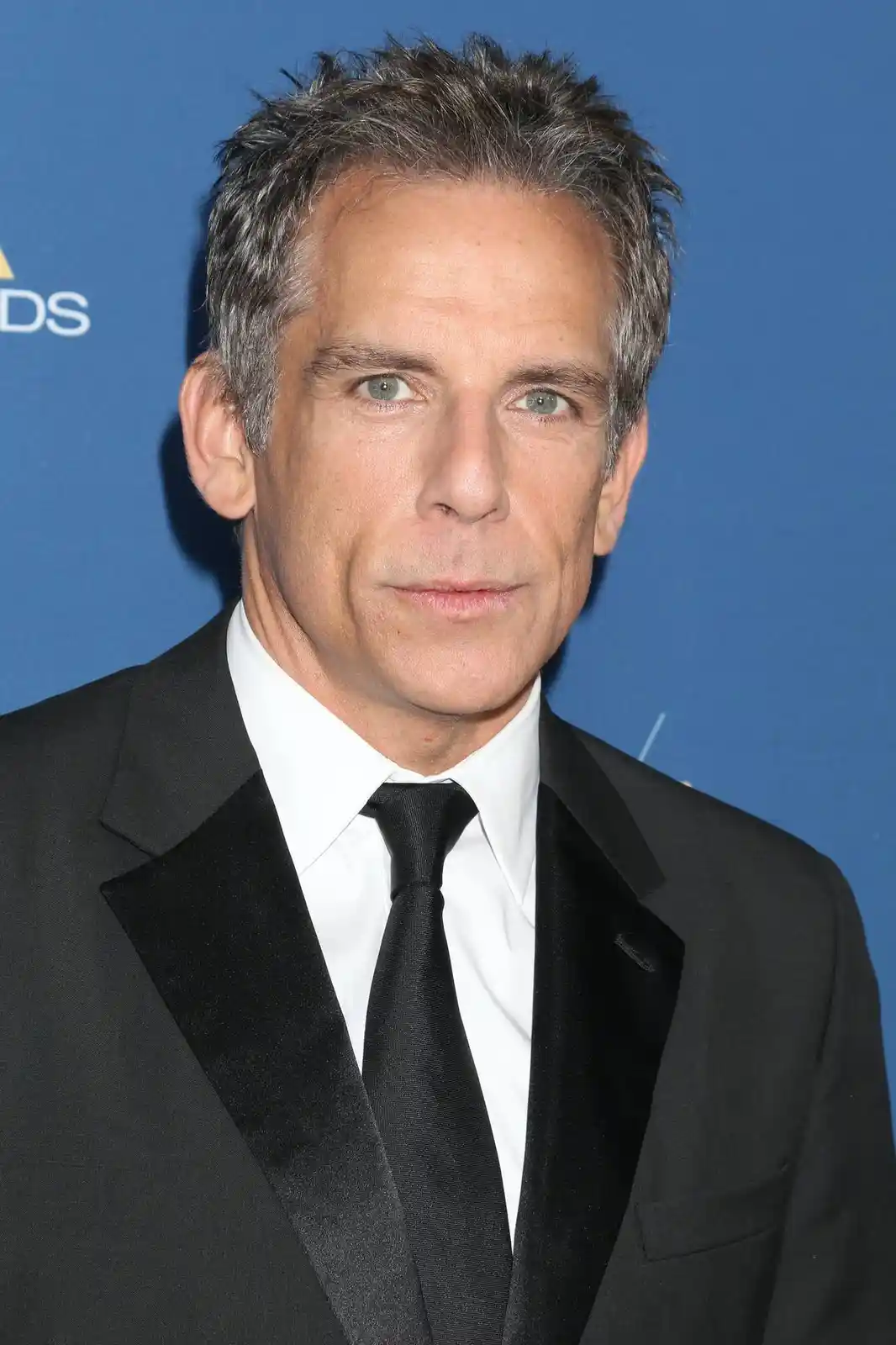 A man with graying hair and green eyes, wearing a black tuxedo with velvet lapels and a black tie, stares directly at the…