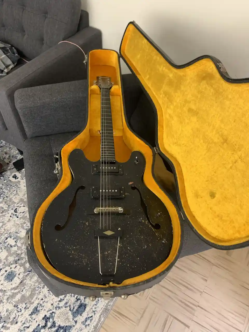 A black electric guitar with a worn, speckled finish sits inside an open, yellow-lined hard case resting on a grey couch…