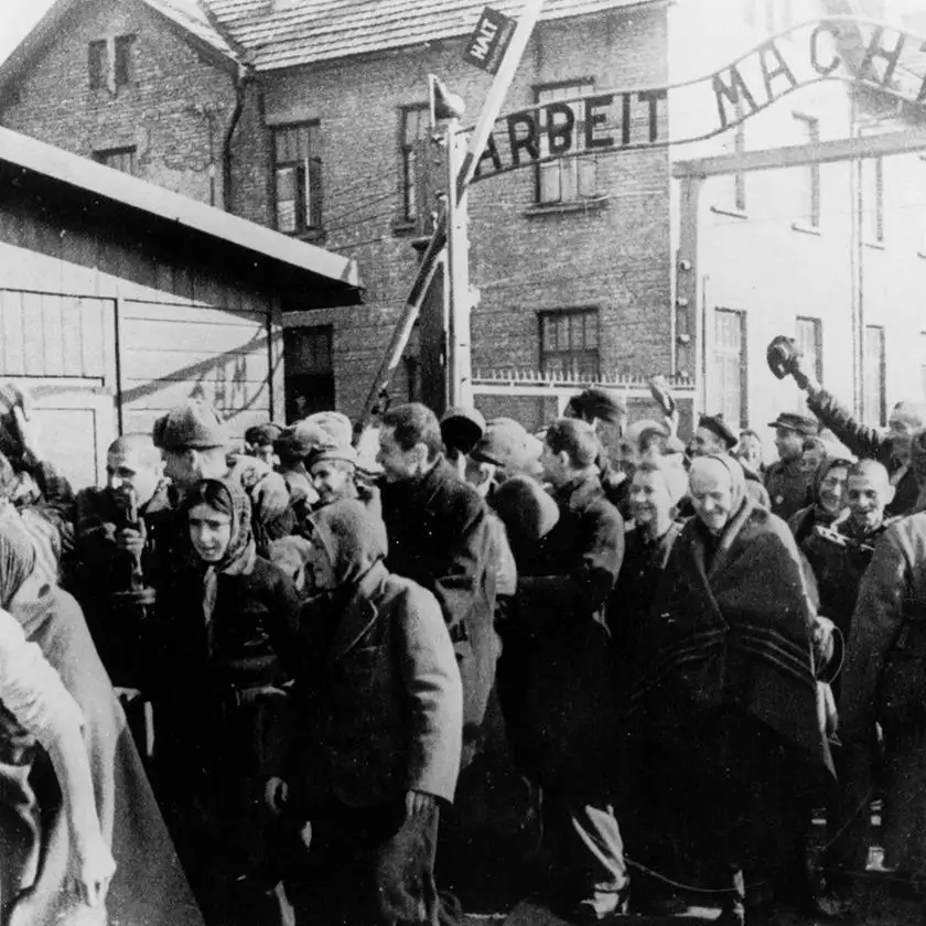 A black-and-white historical photograph shows a crowd of emaciated people, many wearing headscarves or caps, standing before…