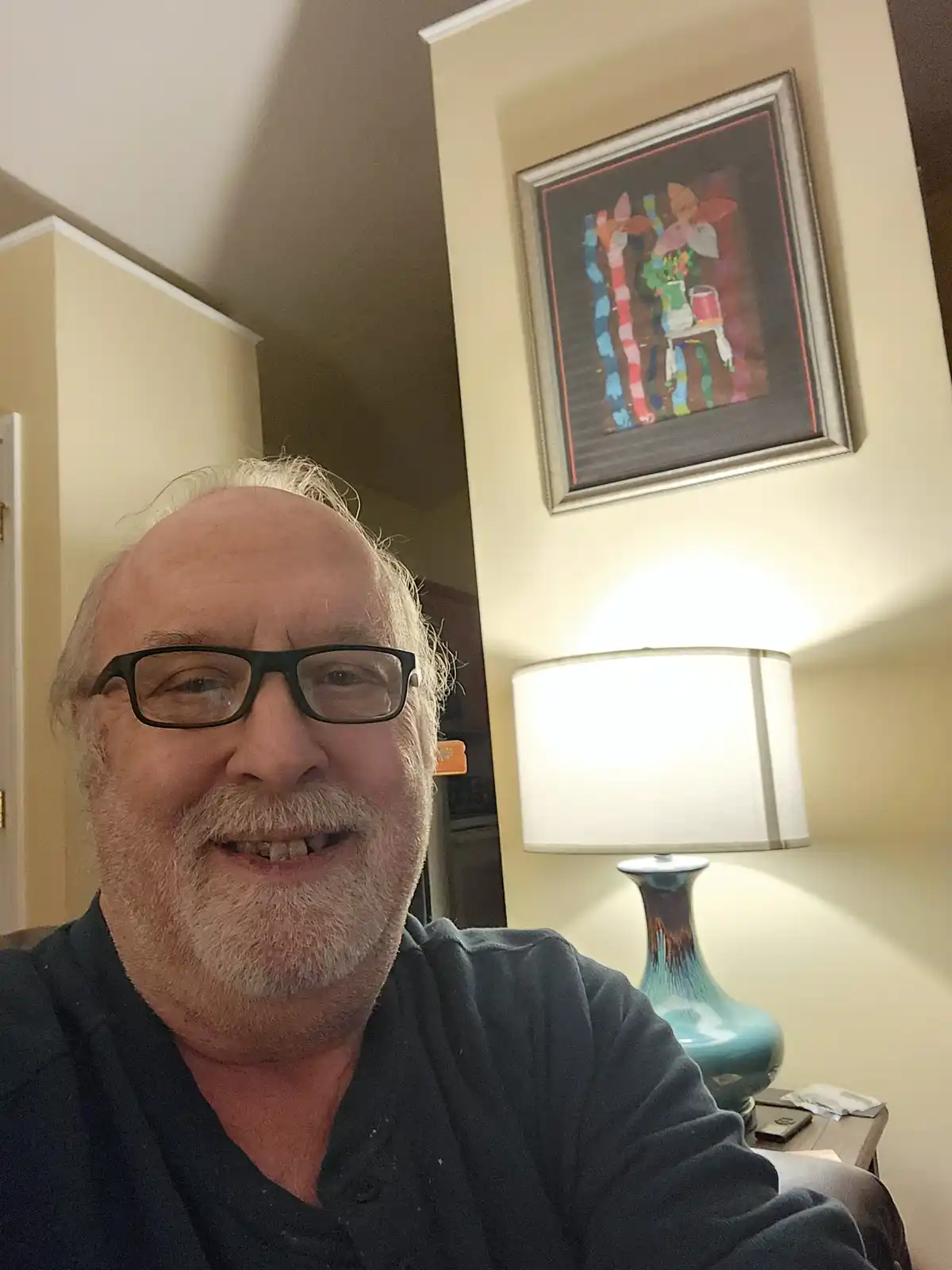 A middle-aged man with glasses and a white beard smiles at the camera while sitting in a room. Behind him, a brightly lit…