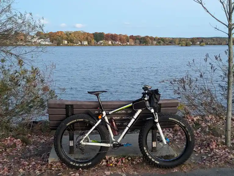 A white fat-tire bicycle is parked on a concrete base next to a dark wooden bench overlooking a calm lake with…