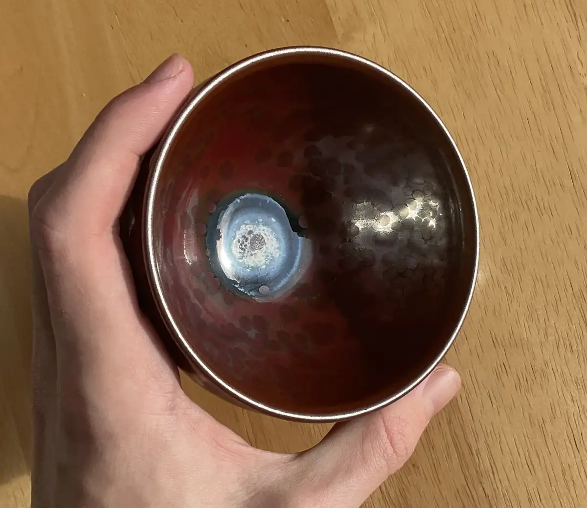 A hand holds a ceramic bowl with a glossy, metallic rim and a rich, dark red interior featuring a swirling, iridescent blue…