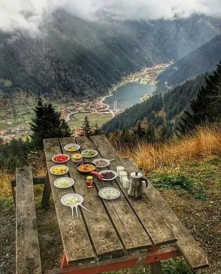 A wooden picnic table set with various dishes and drinks sits on a grassy hillside overlooking a scenic valley with a lake…