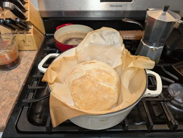 A round loaf of bread with a golden crust sits inside a white Dutch oven, which is lined with brown parchment paper and…