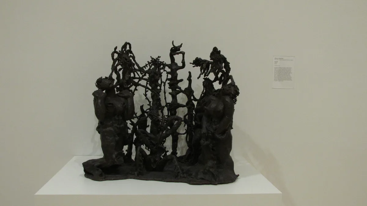 A dark, abstract bronze sculpture with twisted, humanoid forms and tangled branches is displayed on a white pedestal in a…