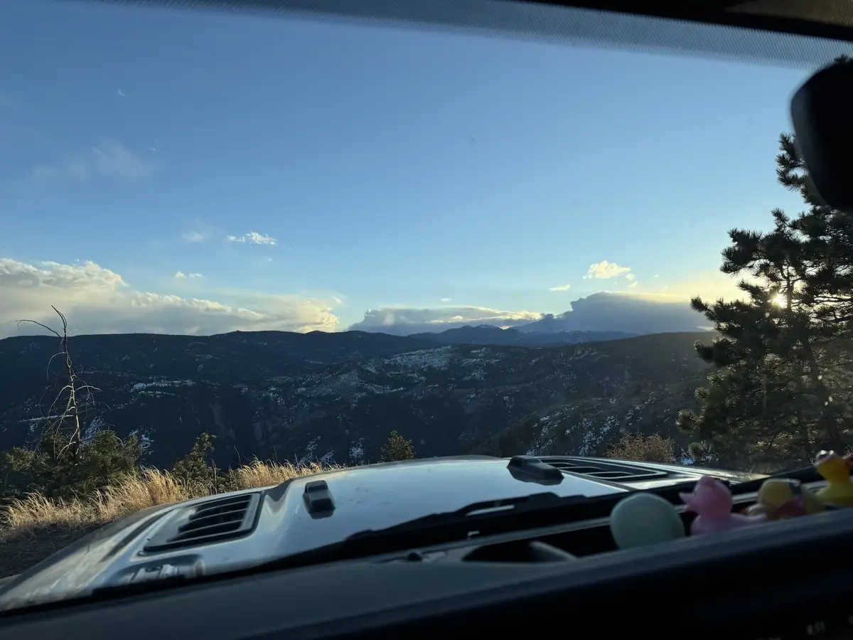 A photo taken from inside a car looking out at a scenic mountain vista during sunset or sunrise, with the car's dashboard…