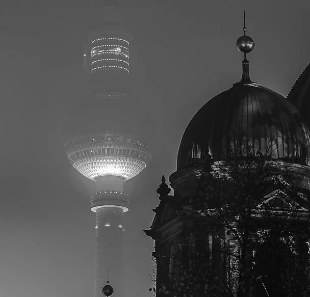 A black and white photograph shows the Berlin TV Tower partially obscured by fog, with the dome of a classical building in…