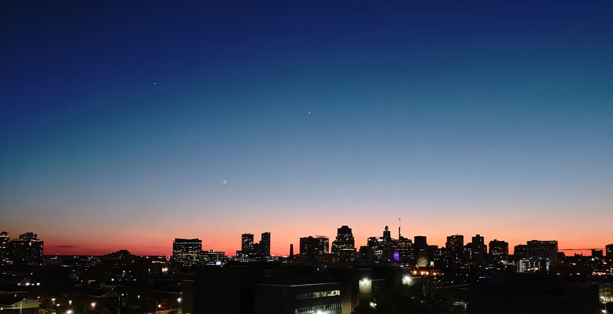 A city skyline at dusk with a gradient sky from orange to deep blue, featuring a crescent moon and two bright stars. The…