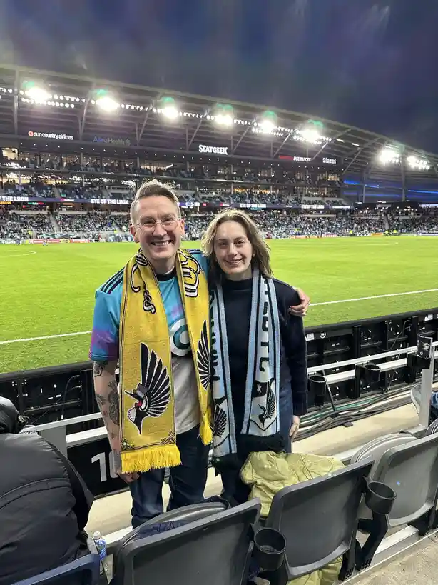 A man and woman pose for a photo at a brightly lit soccer stadium, both wearing team scarves. The man wears a yellow scarf…