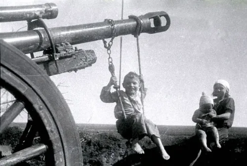 A black-and-white historical photograph shows a child joyfully swinging on a swing suspended by chains from a large…