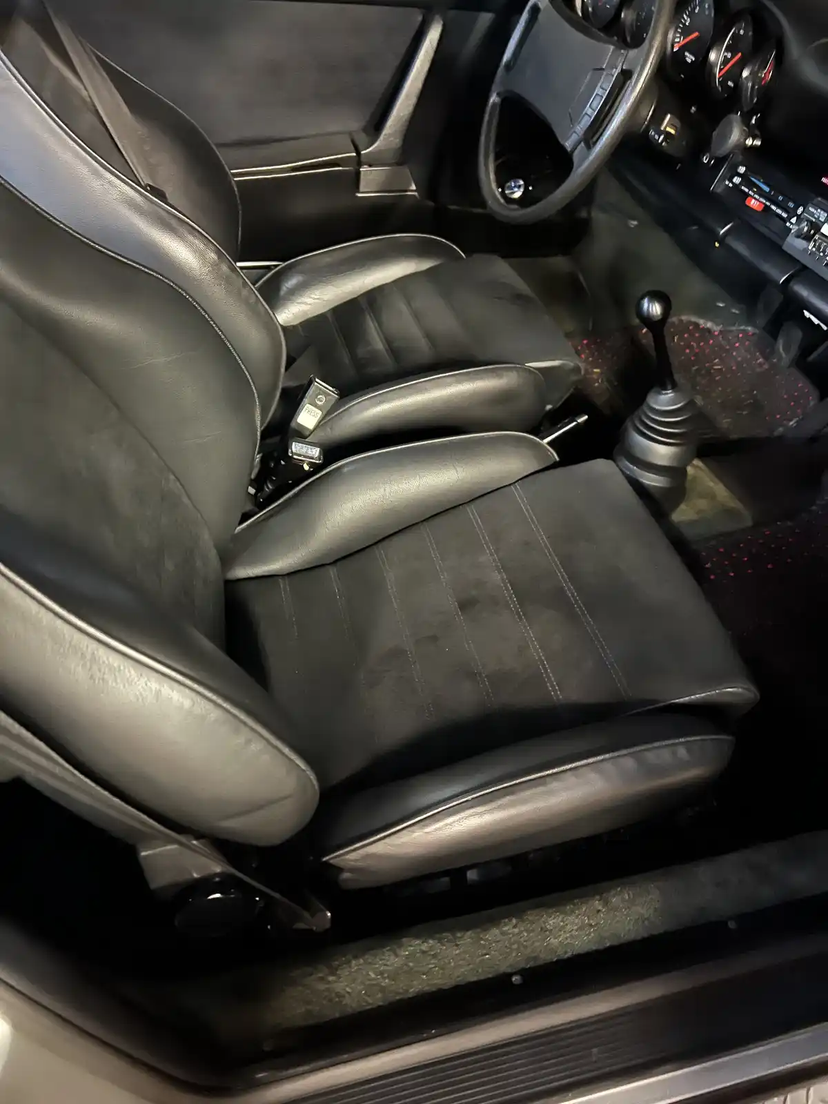 The image shows the interior of a vintage car, focusing on the driver's side with black leather seats, a manual gear stick…