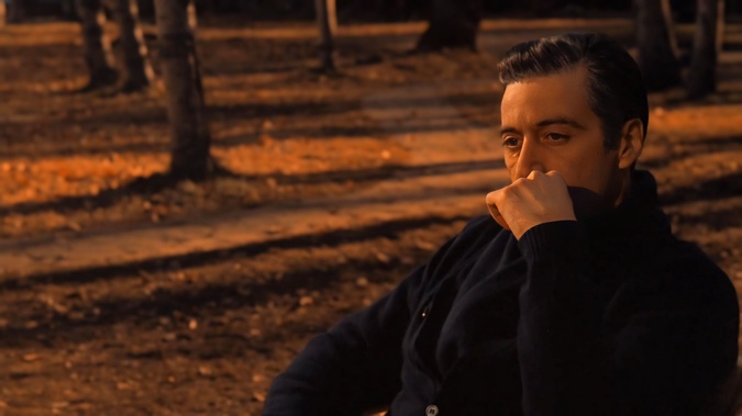 a man with slicked-back hair sits in a park at dusk, hand covering his mouth in shock or disbelief, as if reacting to…