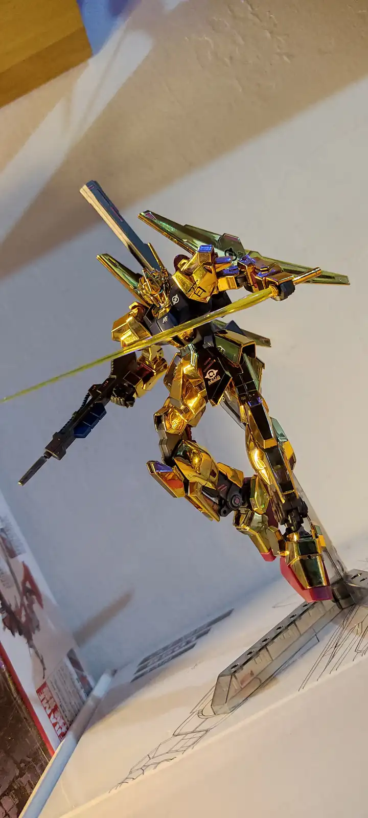 A highly detailed, custom-painted golden Gundam model is posed dynamically on a display stand, holding a sword and rifle…