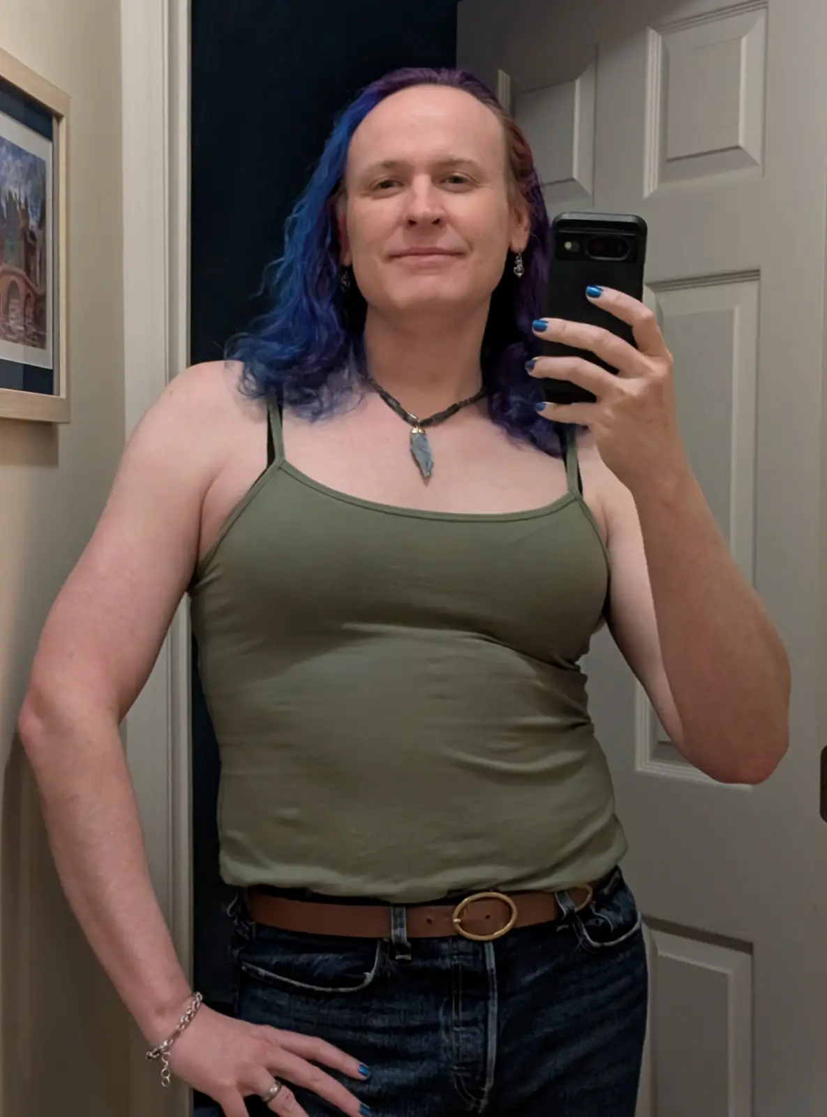 A person with blue and purple hair takes a mirror selfie, wearing an olive green tank top, jeans, and a brown belt with a…