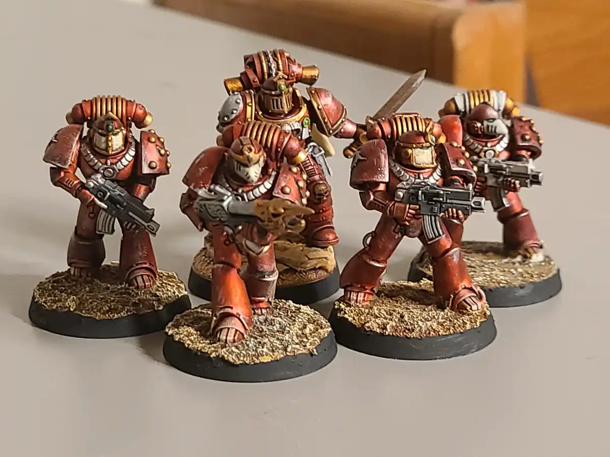 Four intricately painted miniature figures in red and gold power armor stand on textured bases, each holding a weapon. The…