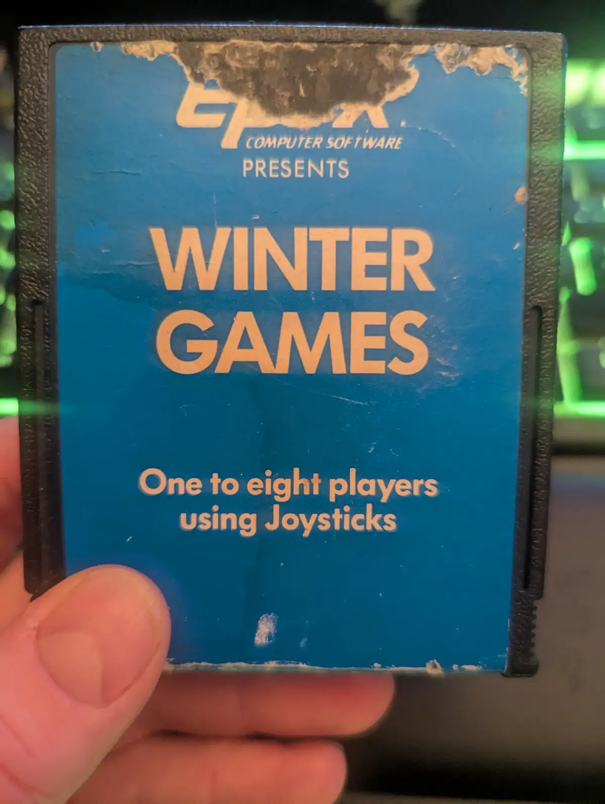 A close-up photo shows a hand holding a heavily worn, blue vintage computer game cartridge. The label, partially torn at the…