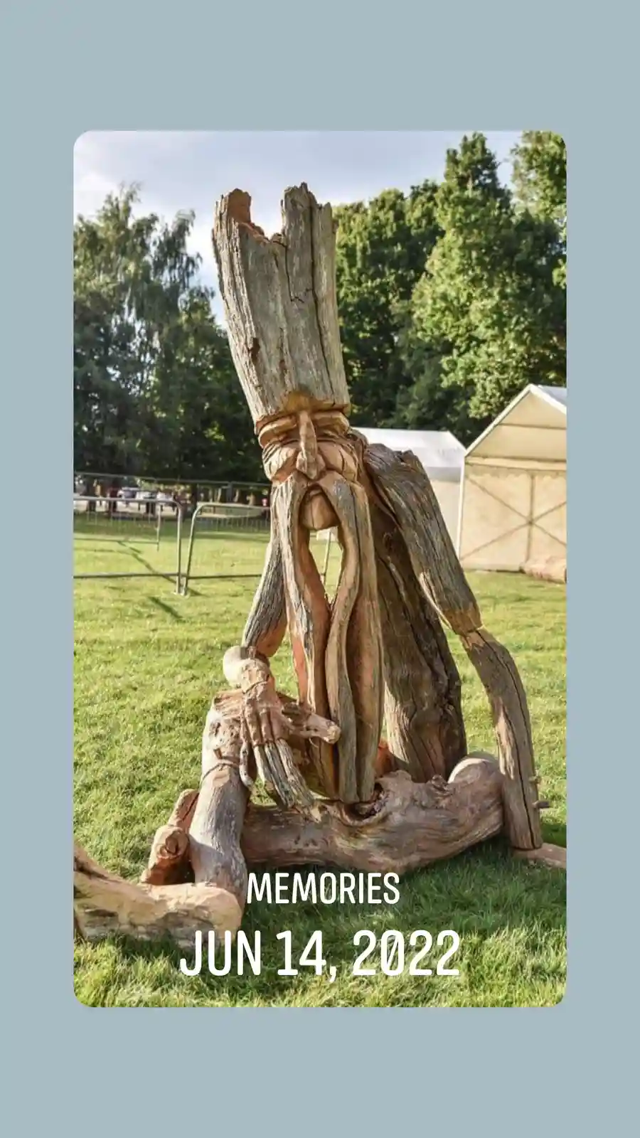A wooden sculpture of a seated, bearded figure carved from driftwood sits on grass with trees and a white tent in the…