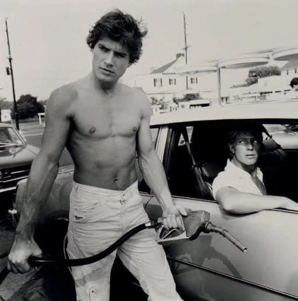 a black-and-white photo shows a shirtless man with defined abs holding a gas pump nozzle at a vintage car, while another man…