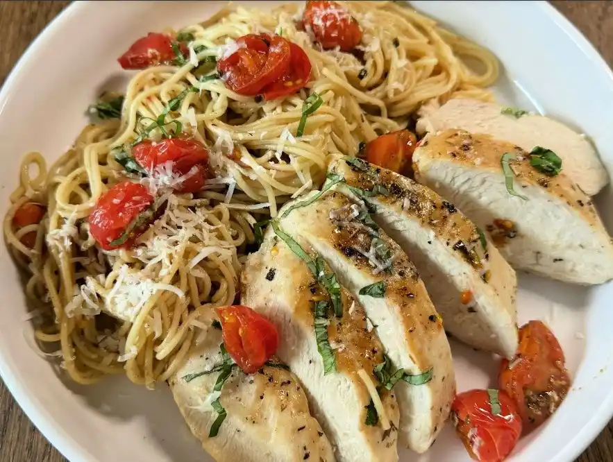 A white plate holds a meal with sliced grilled chicken breast and spaghetti topped with cherry tomatoes, basil, and grated…