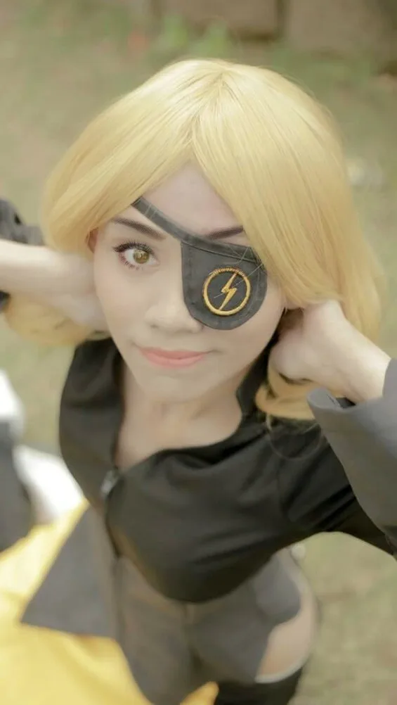 A person with blonde hair and a black eye patch over their left eye, which features a yellow lightning bolt logo, is posing…
