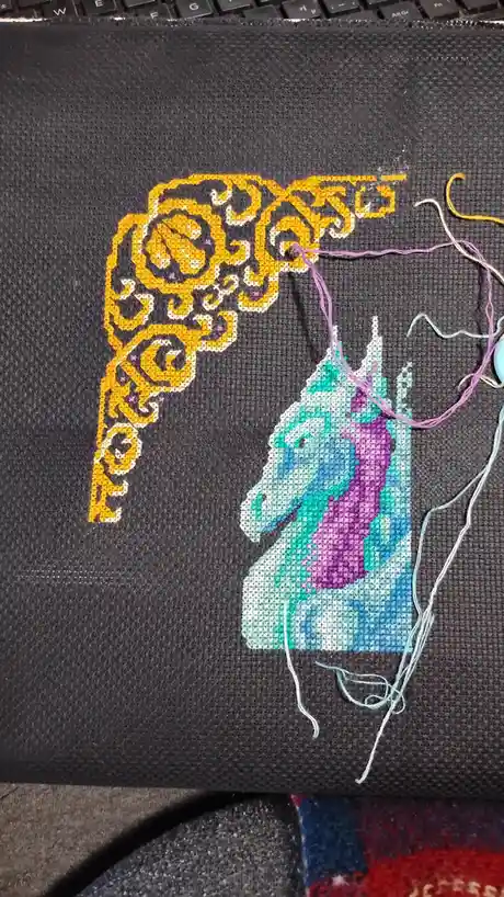 A close-up of a cross-stitch project on dark gray fabric, showing a partially completed unicorn head with a purple mane and…