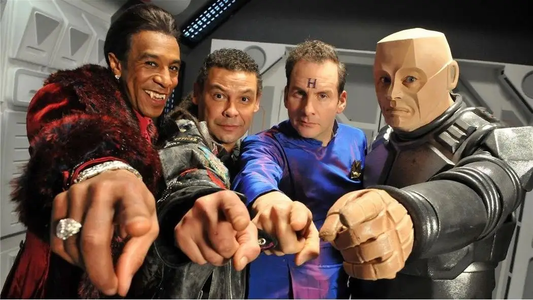 Four men in sci-fi costumes pose with their right fists pointed forward. The man in the center wears a blue uniform with a…