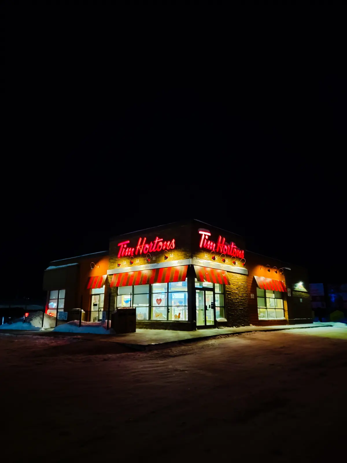 A nighttime photo of a Tim Hortons restaurant illuminated by its bright red neon sign against a dark sky, with snow covering…
