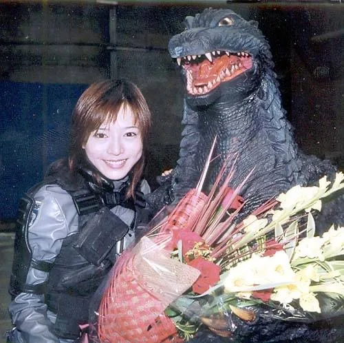 a smiling woman in a black and grey tactical-style outfit stands beside a large, roaring Godzilla costume holding a bouquet…