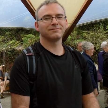 A man with glasses and a black t-shirt stands in front of a modern architectural structure with a curved roof and greenery…