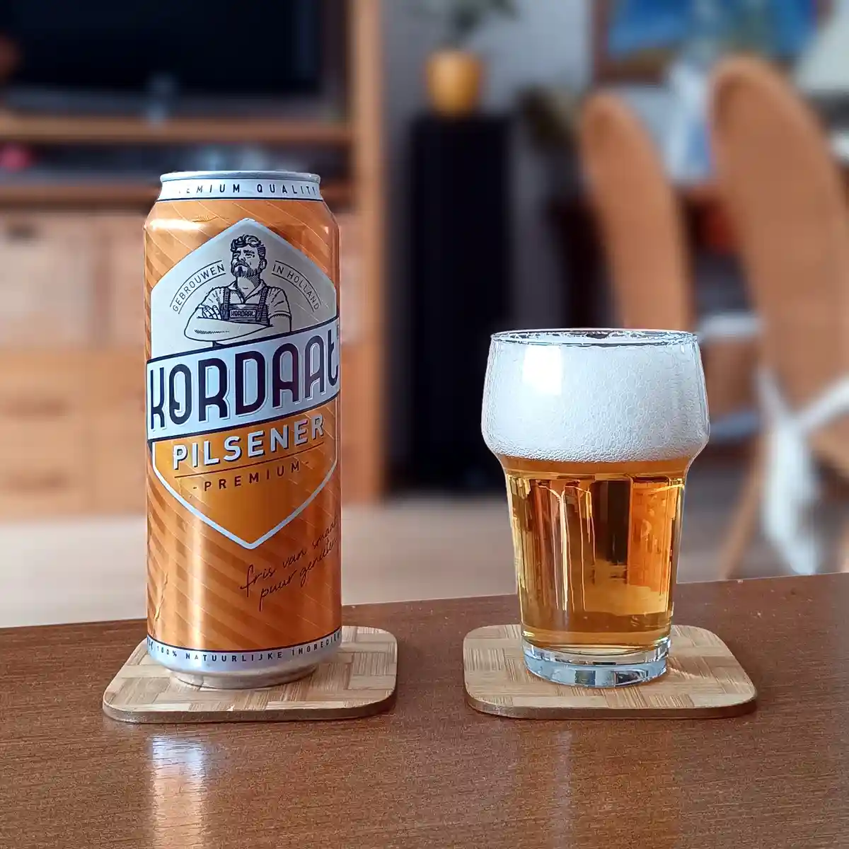 An orange Kordaak Pilsener Premium beer can sits next to a glass filled with golden beer and white foam, both resting on…