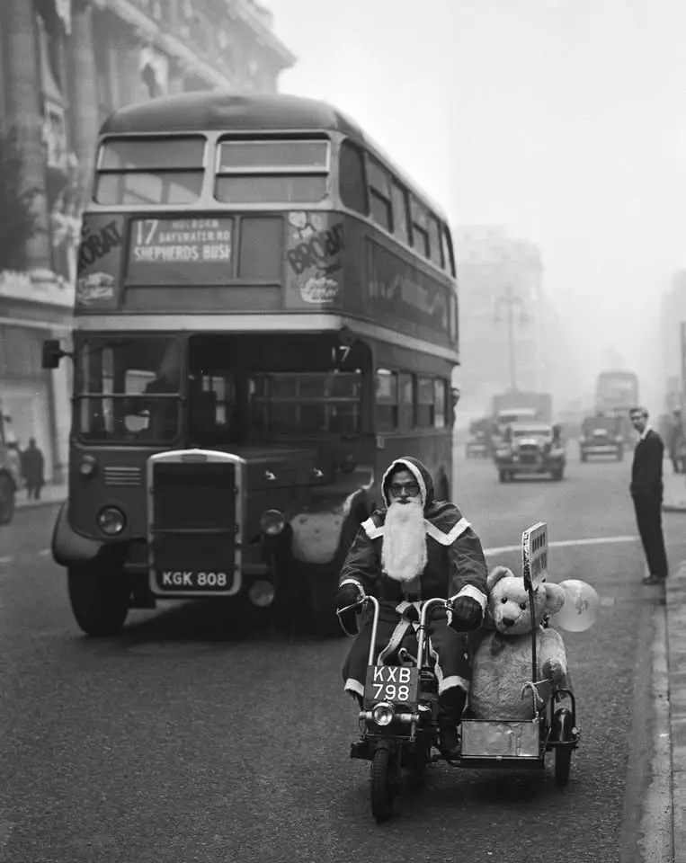 A black-and-white photograph shows a man dressed as Santa Claus riding a moped with a large teddy bear in the sidecar, on a…