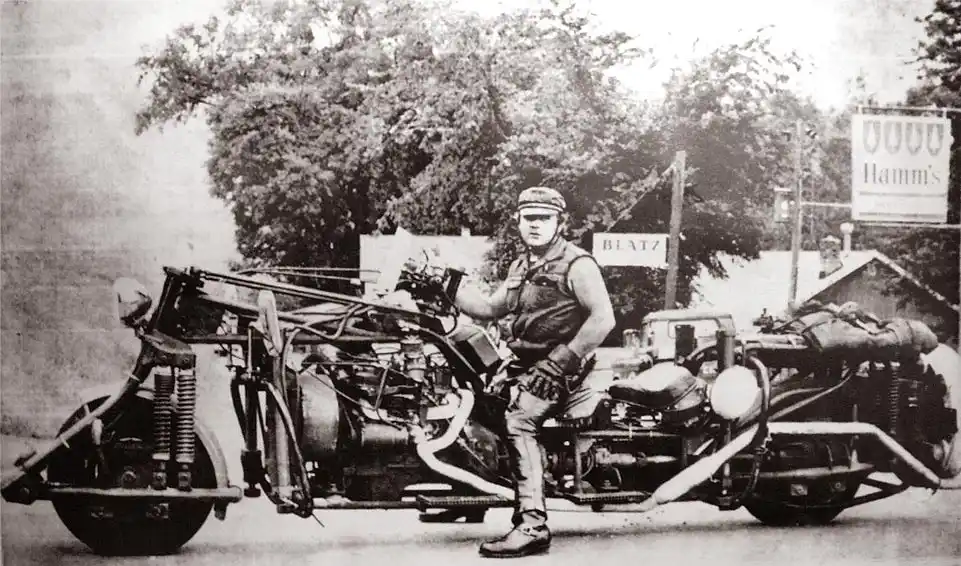 A black-and-white historical photo shows a man in early 20th-century attire sitting on an extremely modified, heavily…