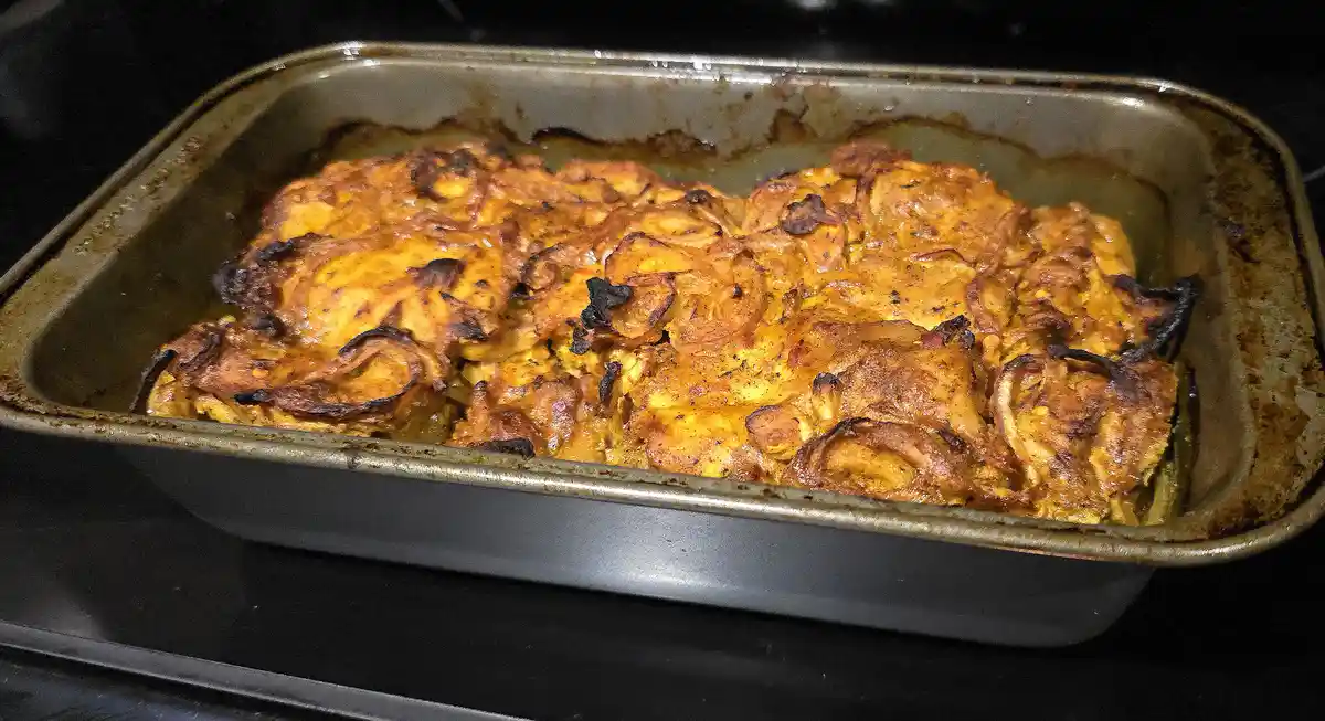 A close-up photo shows a rectangular metal baking dish containing a golden-brown baked dish with caramelized, slightly burnt…