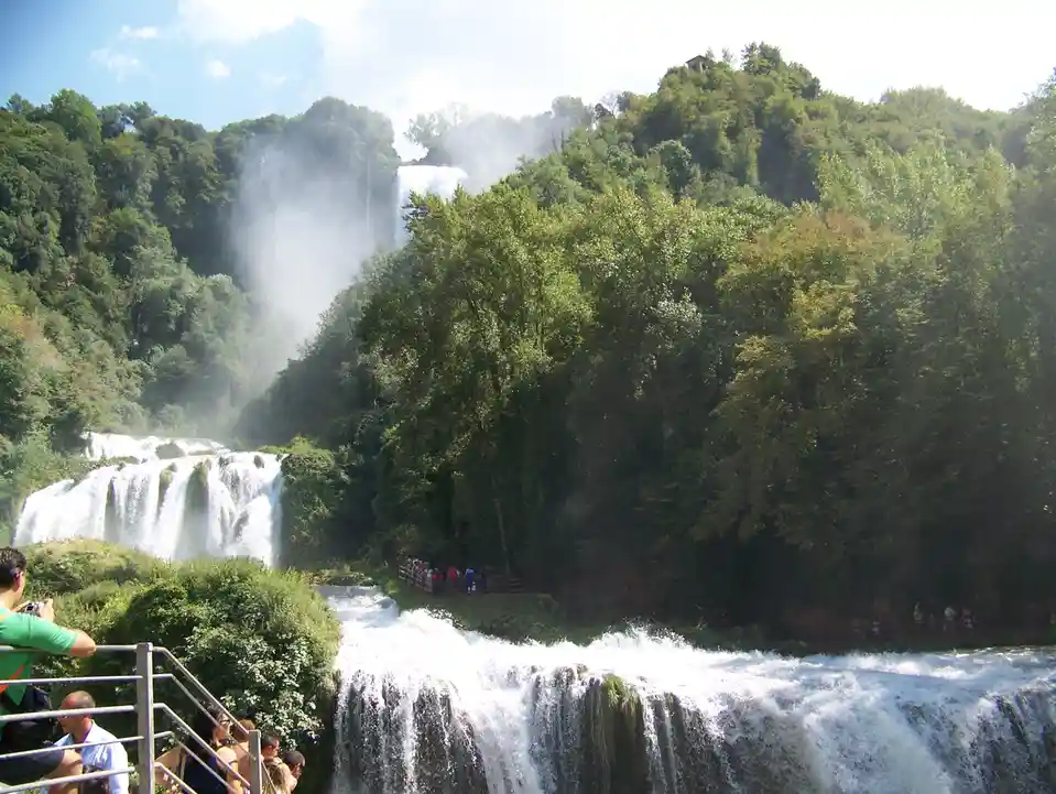 A scenic view of a multi-tiered waterfall surrounded by lush green forests under a partly cloudy sky, with tourists visible…