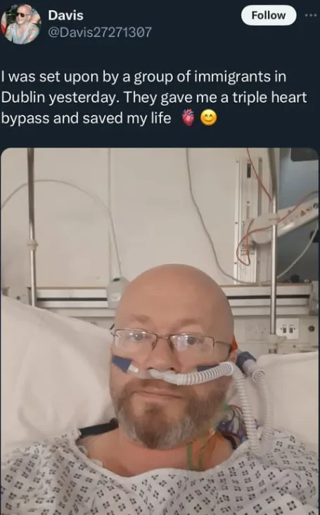 A man with glasses and a beard lies in a hospital bed wearing an oxygen mask and a patterned hospital gown. Medical…