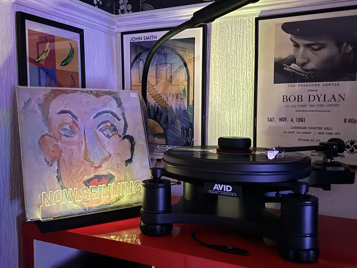 A black AVID Sequel turntable spins a record on a red shelf, with a glowing neon sign reading 'NOW SPINNING' in front of it…