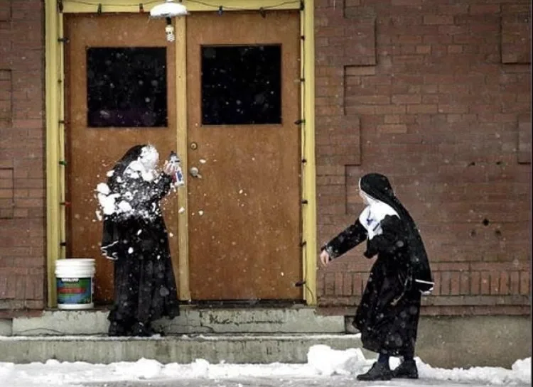 Two nuns in black habits stand outside a brick building with wooden doors, engaged in a snowball fight. One nun is…