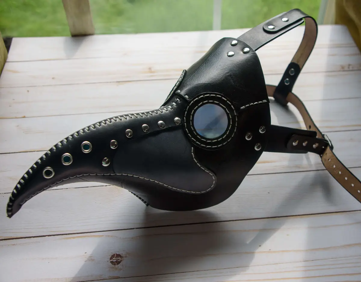 A black leather plague doctor mask with a long beak, silver rivets, and white stitching rests on a white wooden surface. The…