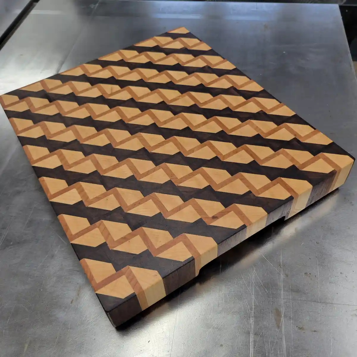A square wooden cutting board with a striking geometric pattern made of alternating light and dark wood pieces arranged in…