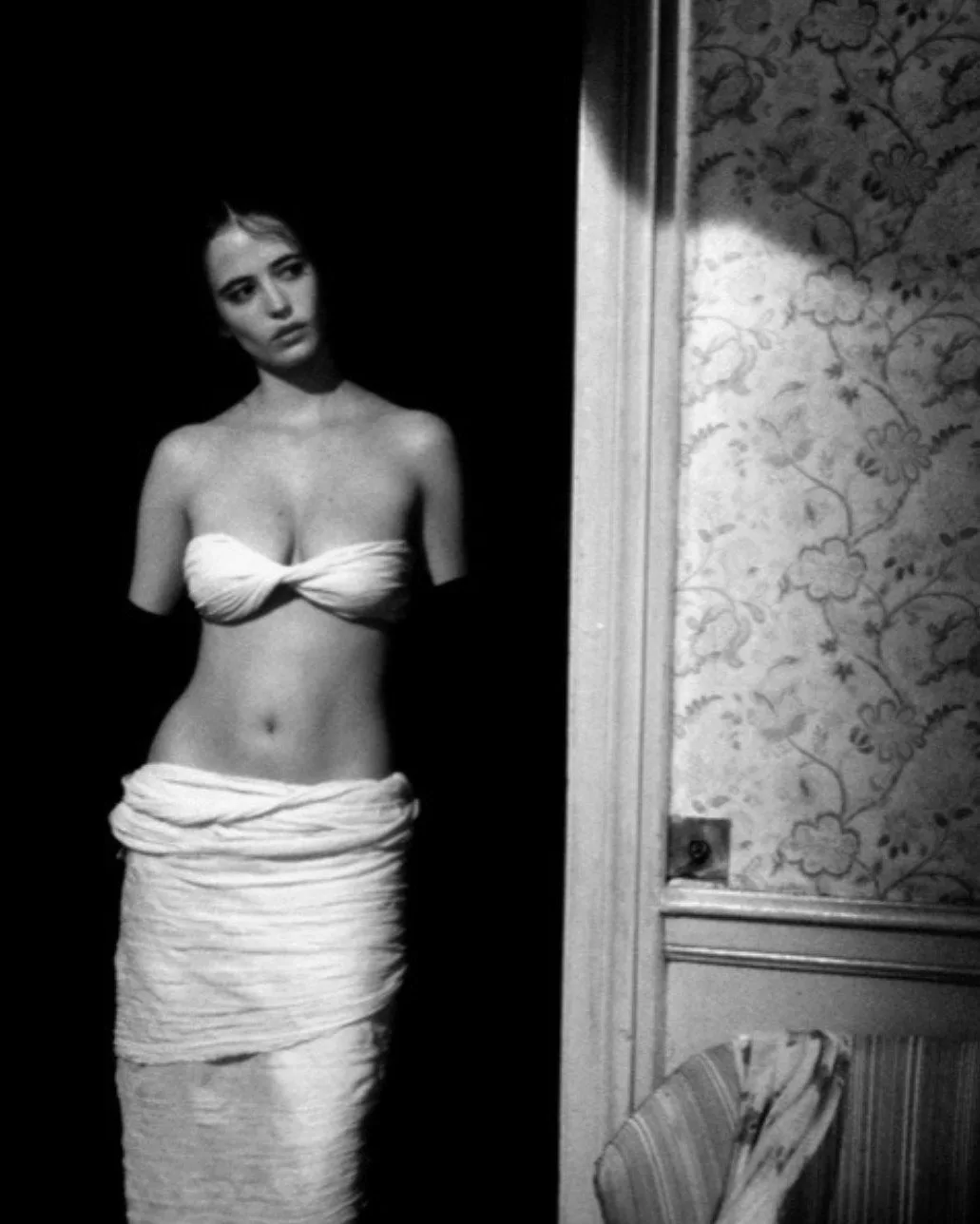 A black-and-white photo shows a woman standing in a dimly lit room, wrapped in a white towel around her waist and chest…